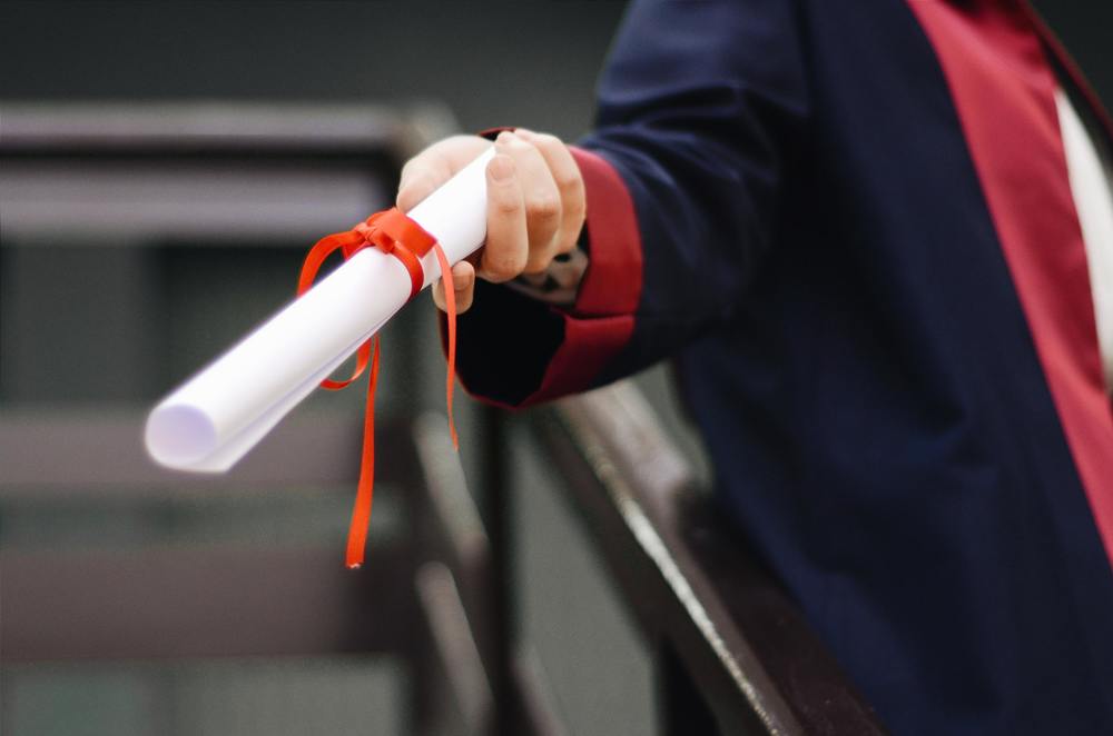 a graduate holding a white diploma tied with a red ribbon in a black gown